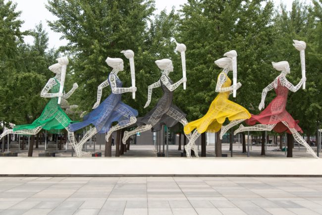 five running women wearing different dresses holding torch statue